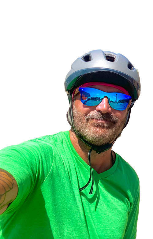 Man with a beard wearing a silver bike helmet, blue reflective sunglasses, and a bright green shirt taking a selfie outdoors.