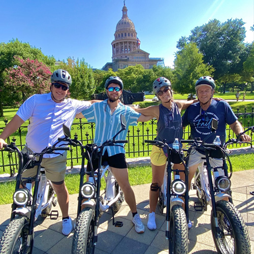 Four people wearing helmets posing with electric bikes in front of a capitol building on a sunny day.