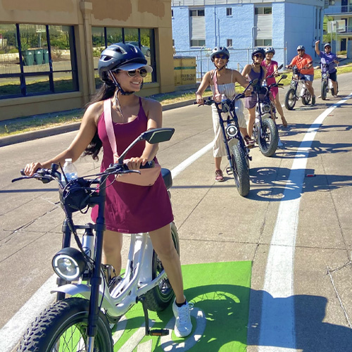 Five people wearing helmets riding electric bikes on a sunny urban street.
