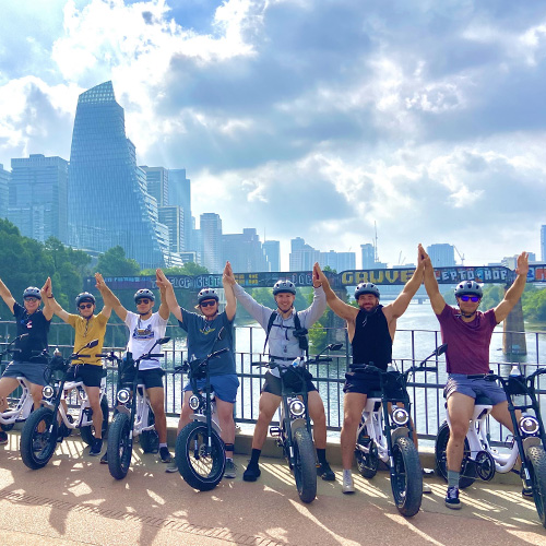 Seven people on electric bikes with helmets high-fiving in front of a river and city skyline under a blue sky with clouds.