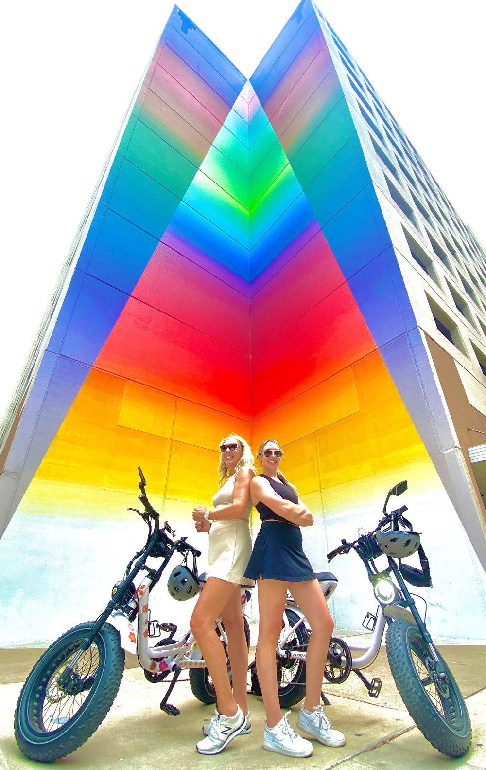 Two women standing back to back with electric bikes in front of a tall building corner painted with a vibrant rainbow gradient mural.
