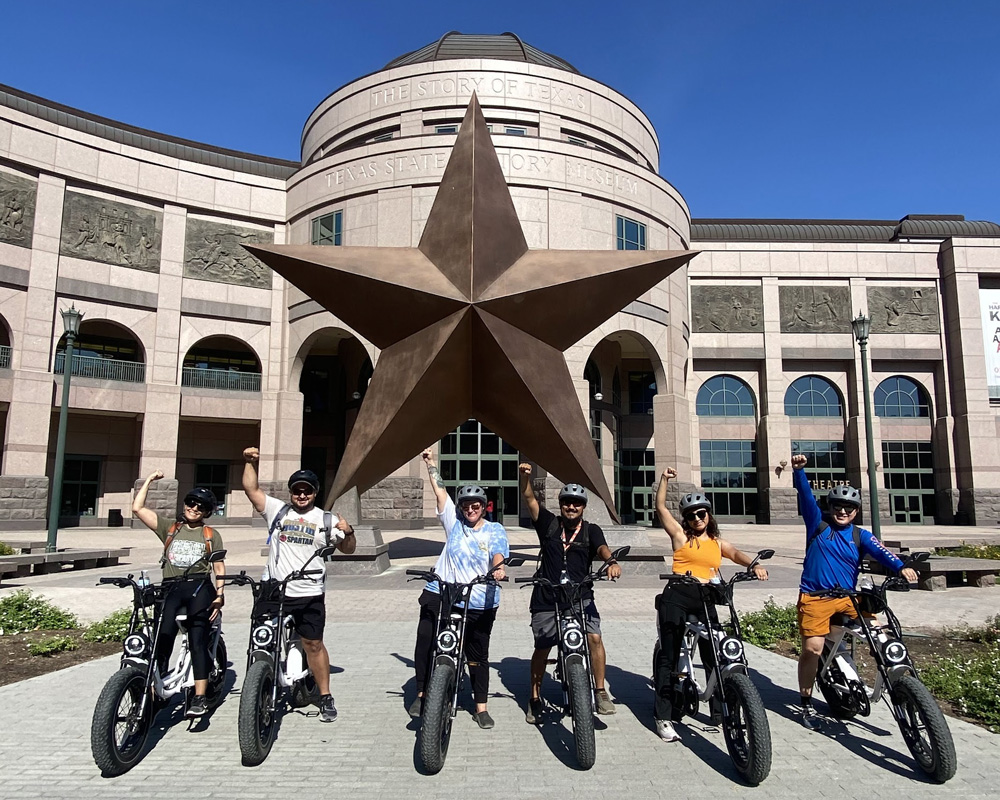 Six people on electric bikes posing with raised fists in front of a large bronze star sculpture at the Texas State History Museum under clear blue sky.