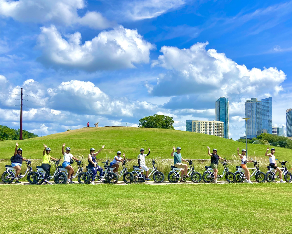 Group of eleven people wearing helmets riding electric bicycles and waving in a park with green grass and city buildings in the background under a blue sky with scattered clouds.