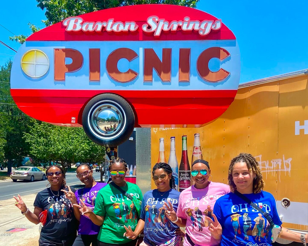 Group of six women wearing colorful matching OHSP T-shirts posing and smiling under a bright red 'Barton Springs Picnic' sign on a sunny day.
