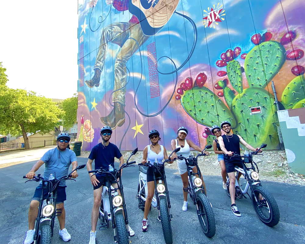 Five people wearing helmets sitting on electric bikes in front of a colorful mural featuring a person playing guitar and cactus plants.
