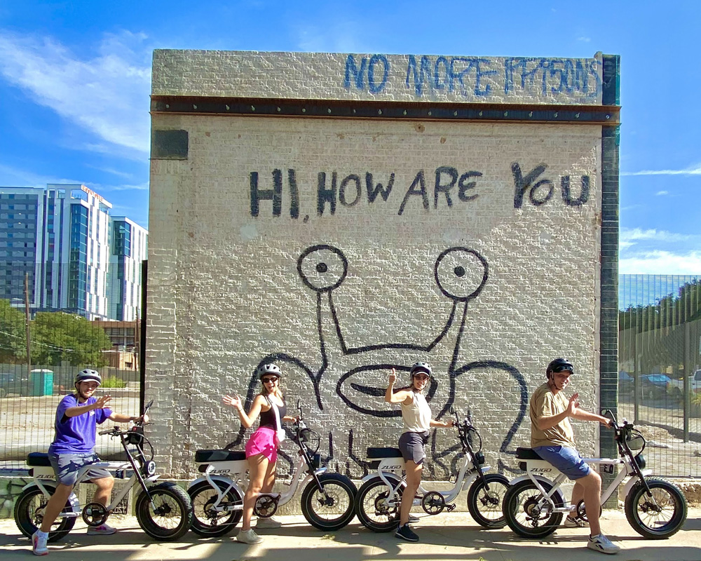 Four people wearing helmets posing on electric bikes in front of a brick wall with graffiti saying 'HI, HOW ARE YOU' and stylized eyes.