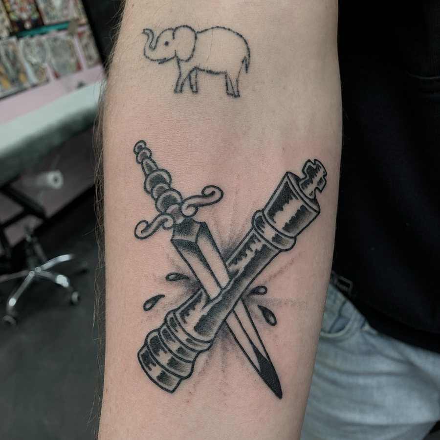 Small elephant above crossed objects in a black-and-grey arm tattoo