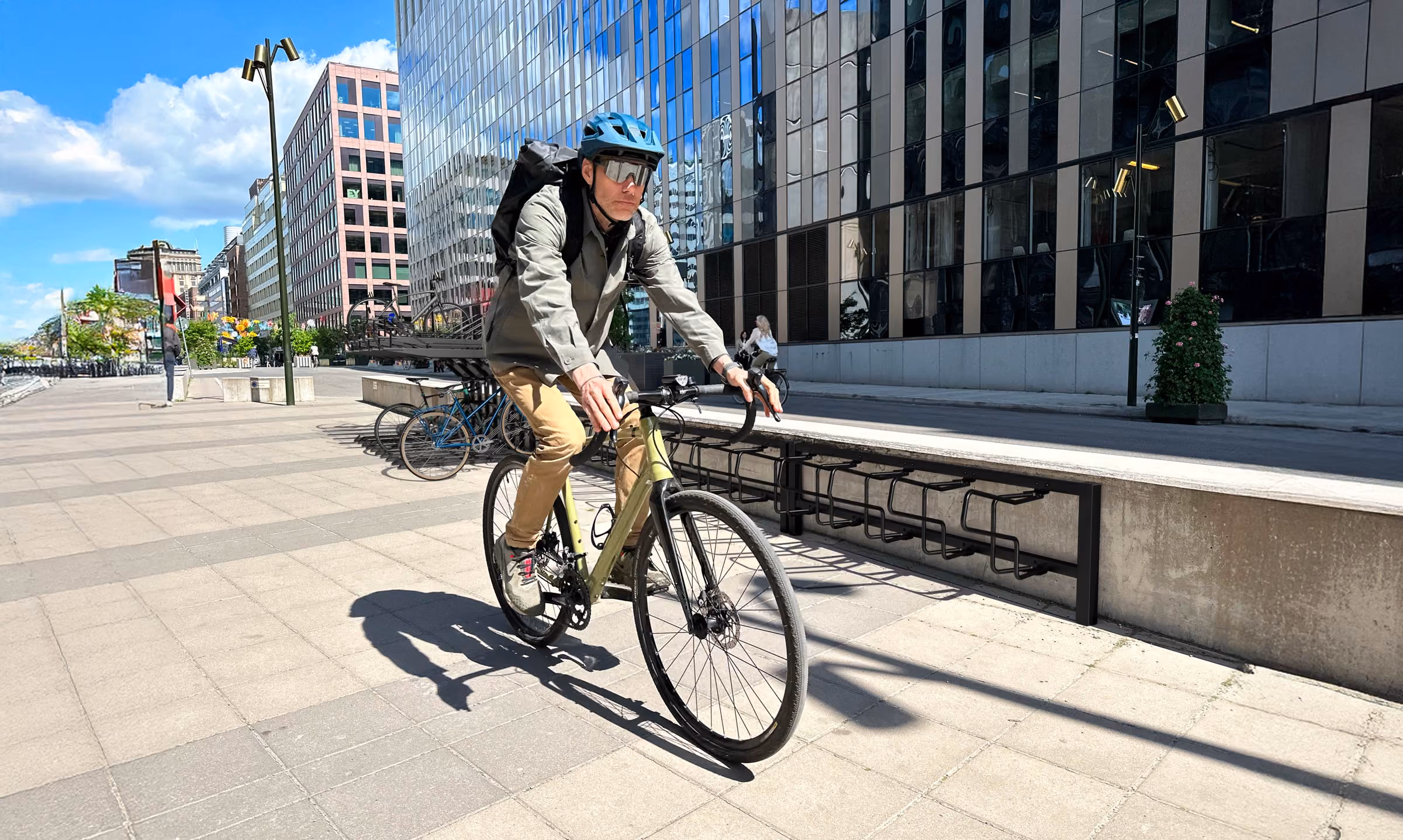 En man med blå cykelhjälm och ryggsäck cyklar på en stadsgata med moderna kontorsbyggnader i bakgrunden.