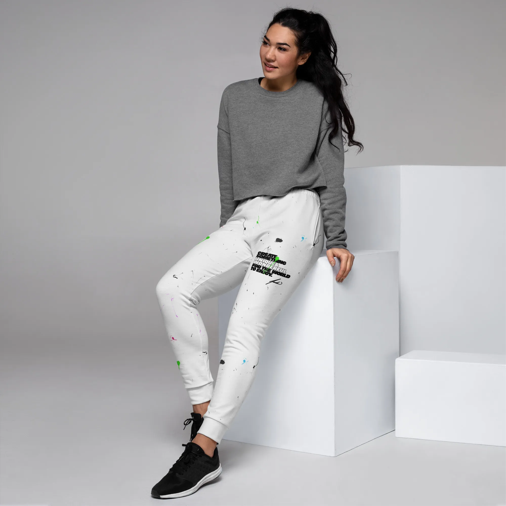 A woman wearing white joggers, with a front, left-side view, showcasing Andrew Hatfield logo, mission statement, and a paint splatter pattern with white, pink, blue, and green colors.