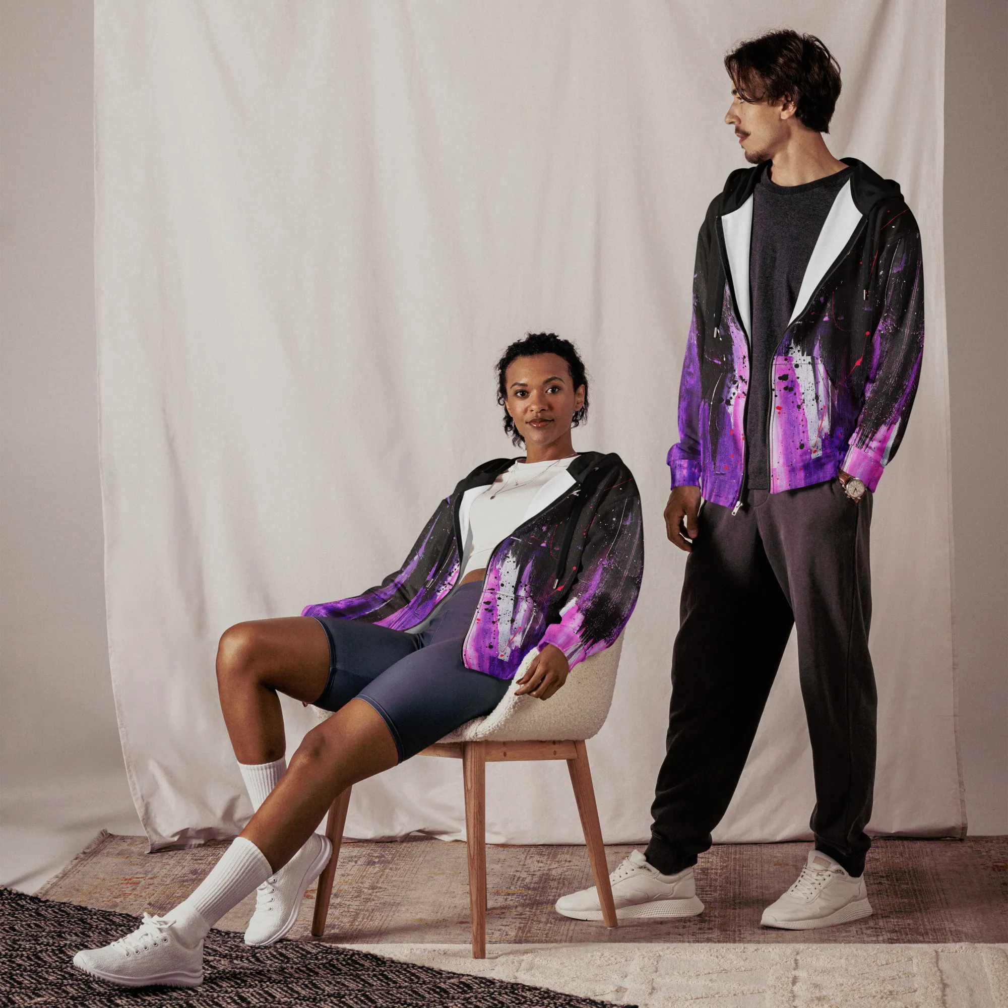 A woman sitting and a man standing wearing a zip hoodie, half zipped, with a front view, showcasing the Andrew Hatfield logo and artwork inspired by 'Dreambound' original painting.