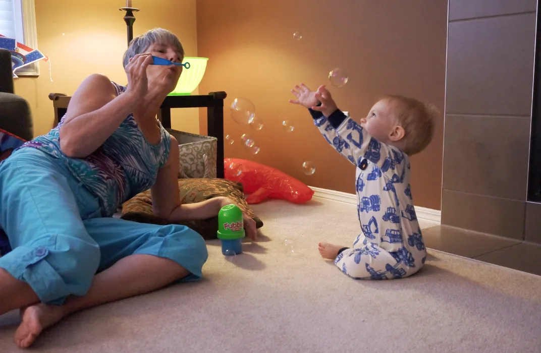 Mary Sampson at 60 years old sits on a living room floor, blowing bubbles for her grandson, as he tries to catch them.