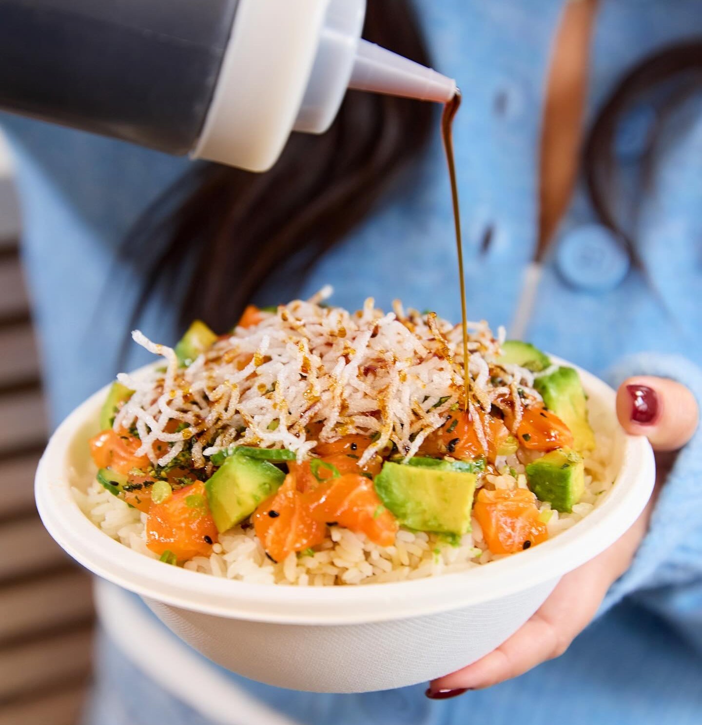 Salmon poke bowl with rice, avocado, toppings, and a sauce being poured on top.