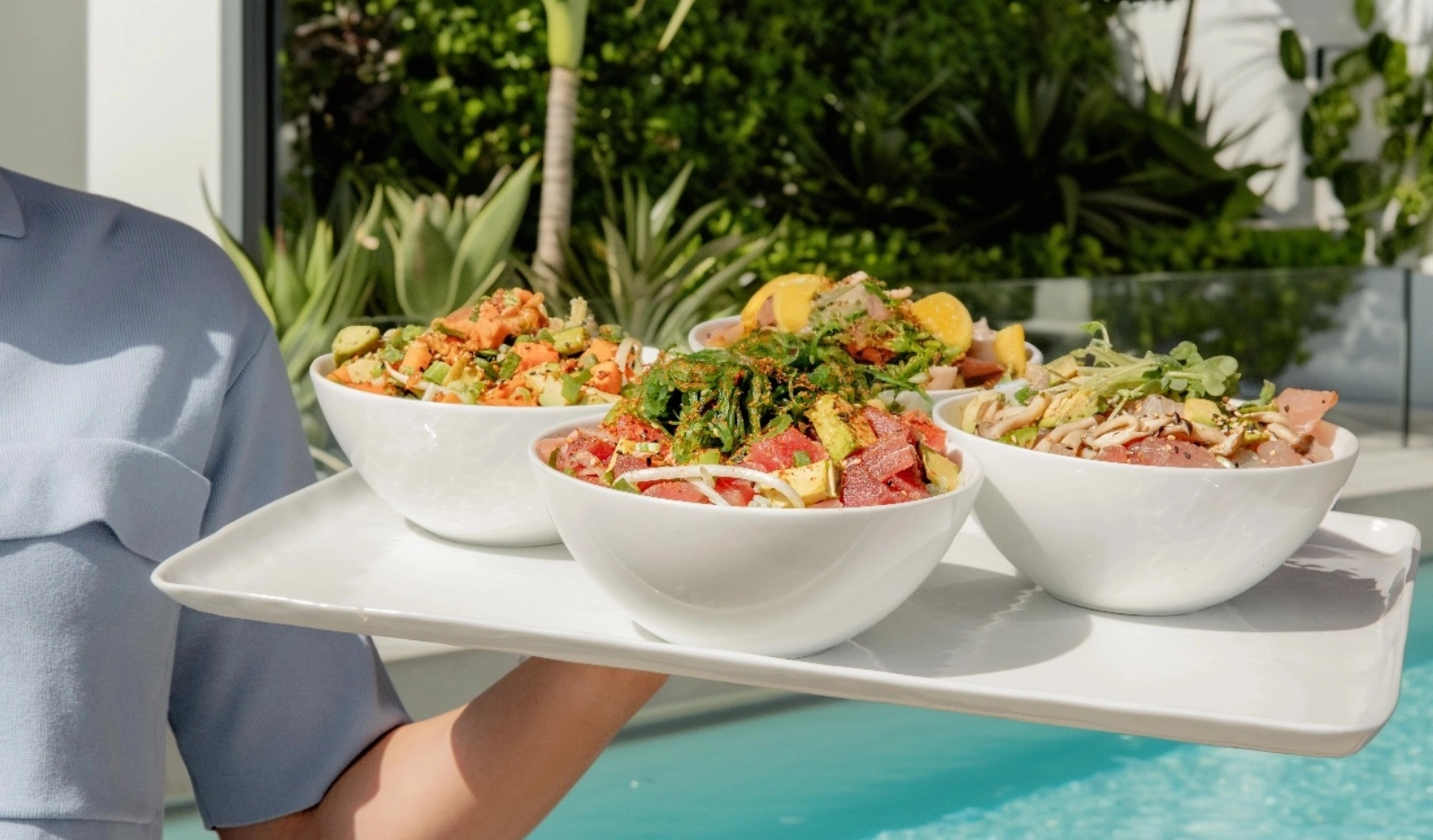 A server carries a tray of colorful Sweetfin poke bowls topped with fresh seafood, avocado, vegetables, and garnishes beside a sunny poolside setting.