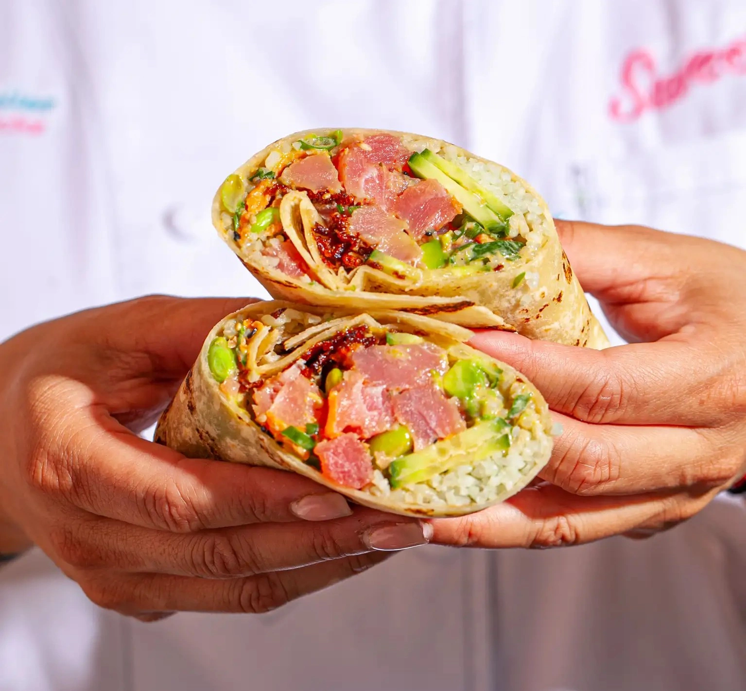 Sweetfin poke burrito cut in half with fresh tuna, avocado, edamame, cucumber, and rice, held by a chef for serving.