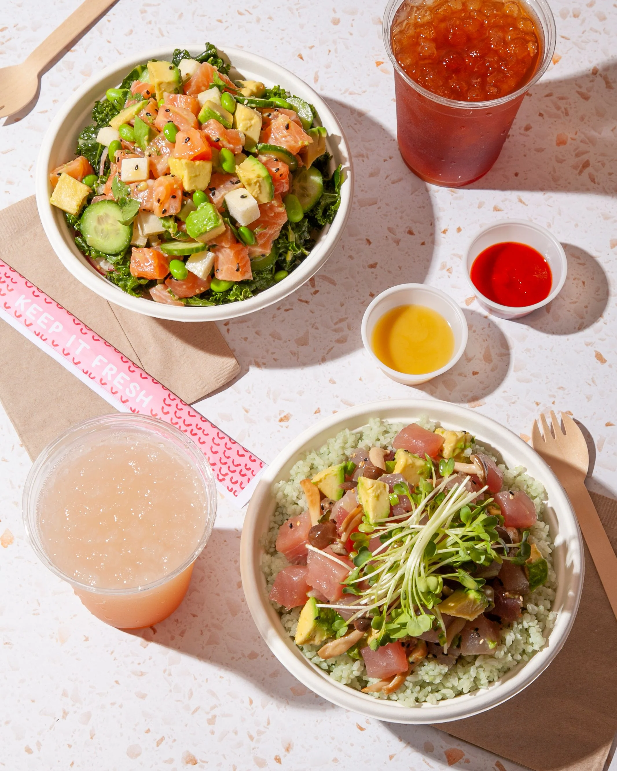 Two Sweetfin poke bowls with fresh vegetables, greens, drinks, and sauces on an outdoor table.