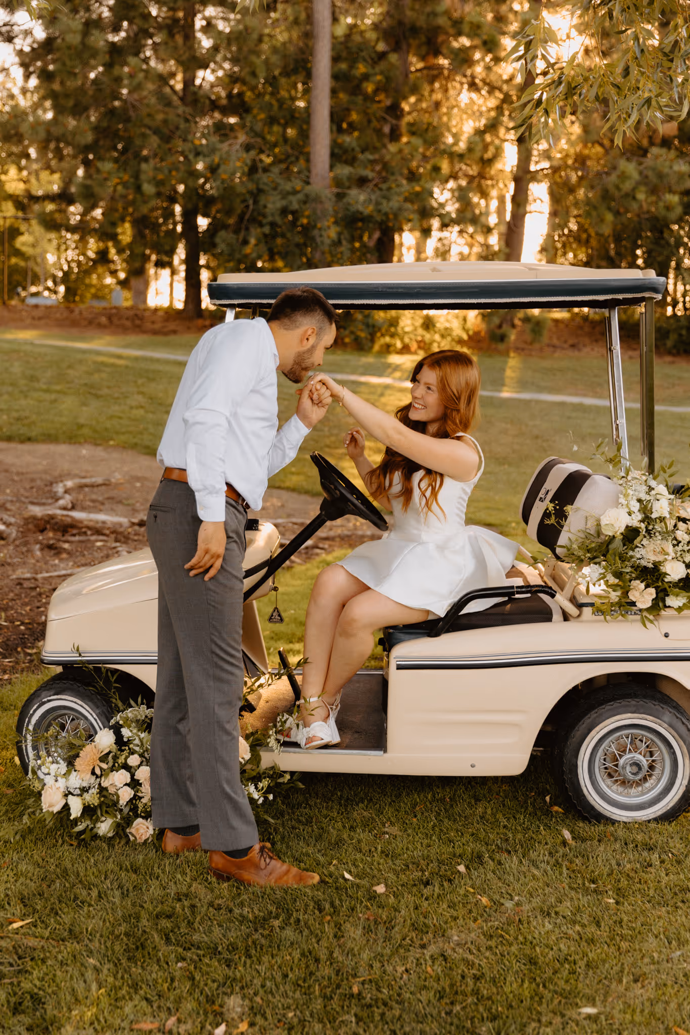 Elegant photo of young engaged couple with vintage golf cart. Groom is kissing hand of the bride while the couple is dressed in classy engagement style outfits.