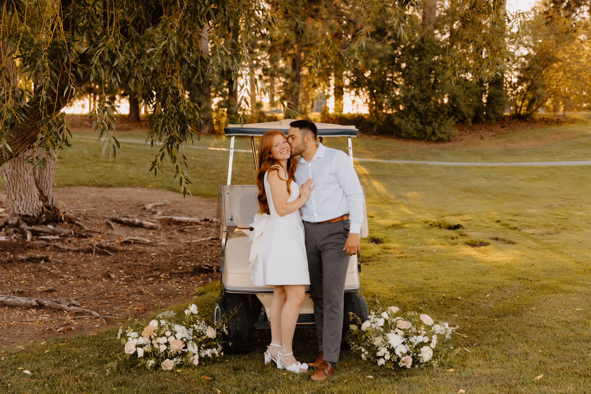 Elegant photo of young engaged couple with vintage golf cart. Groom is kissing the bride while the couple is dressed in classy engagement style outfits.