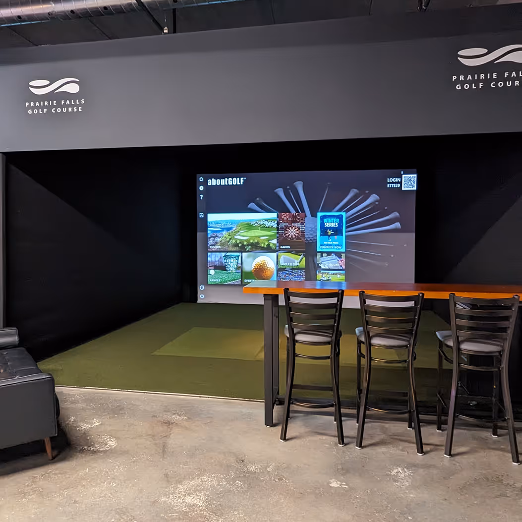 Photo of a virtual golf simulator bay with high top bar table and 3 chairs at the edge of the turf inside the bay. Screen is on showing the simulator software active.