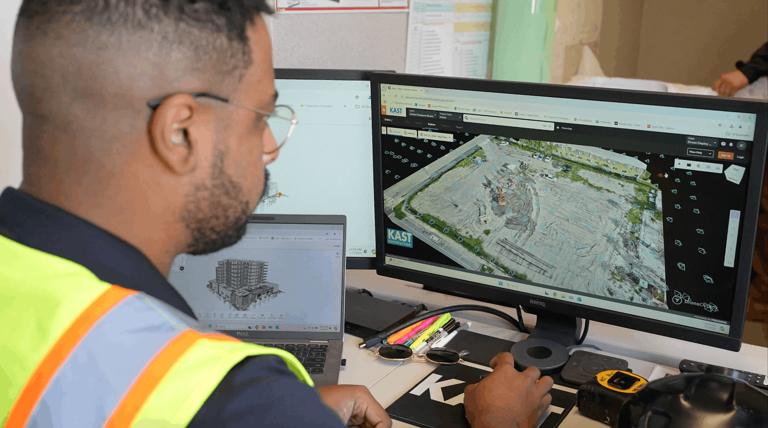 The image depicts a man wearing a high-visibility safety vest, sitting at a desk and working on a co...