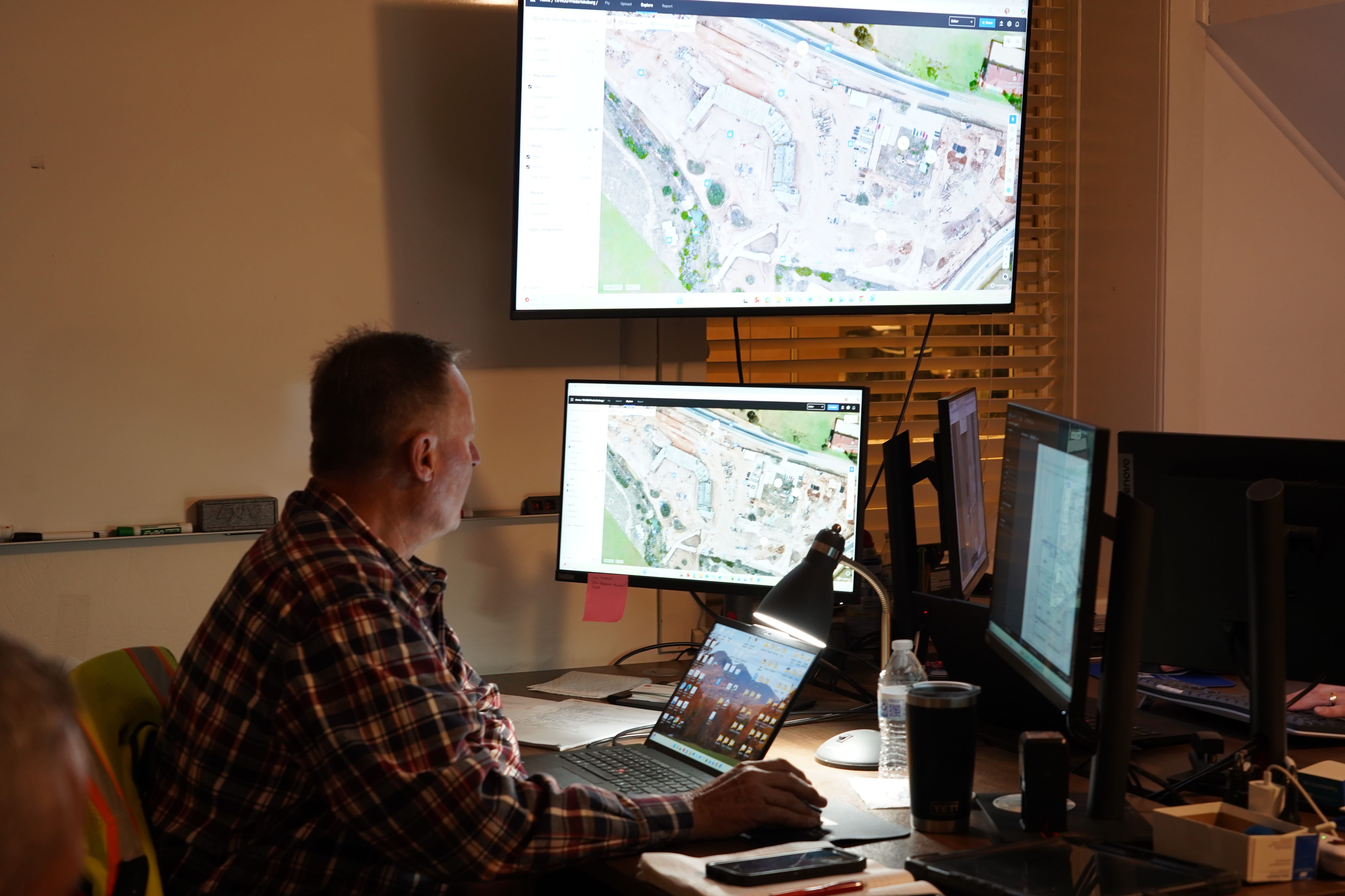A man wearing a brown and orange plaid shirt sits at a desk, viewed from behind, working in what app...