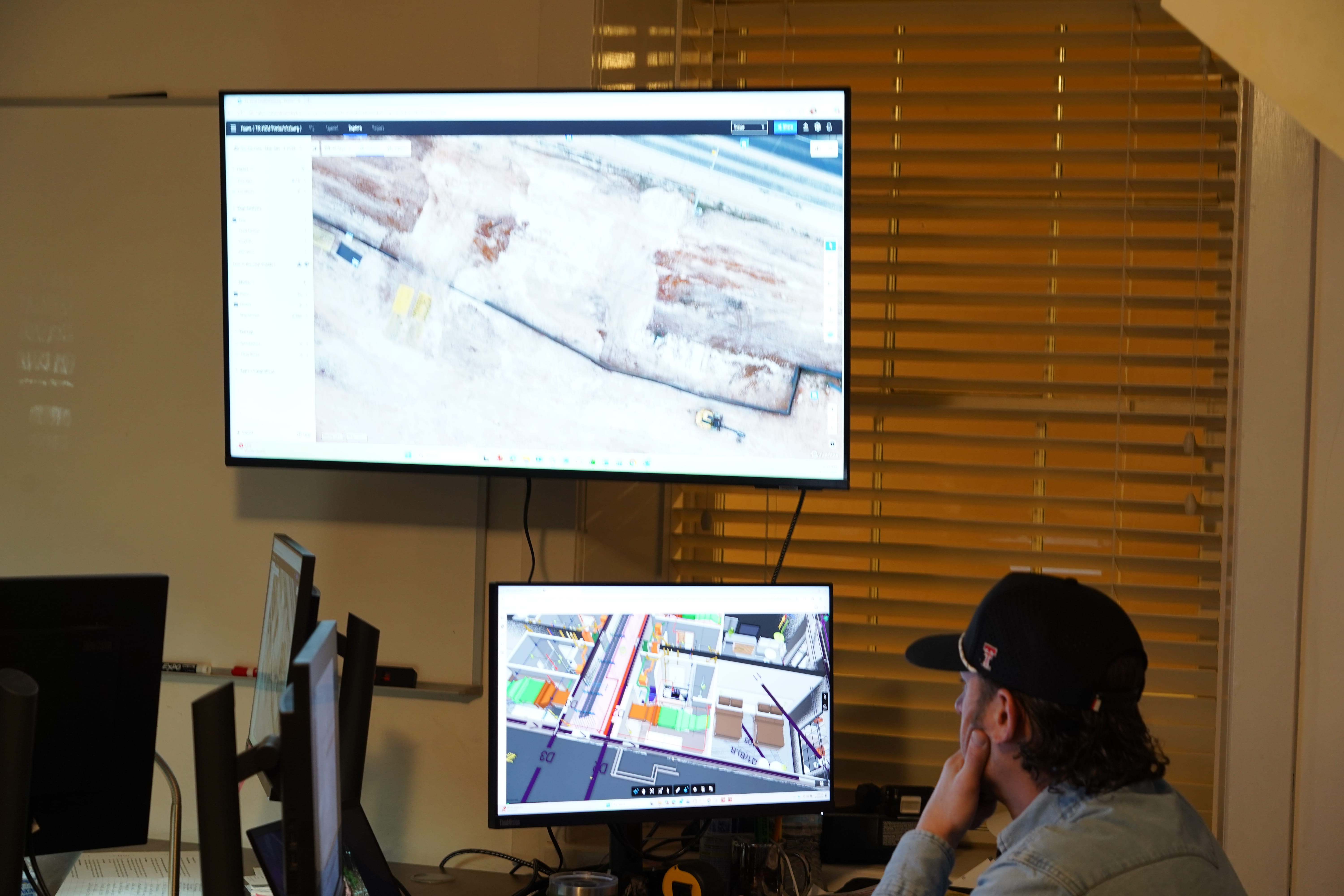 A person wearing a dark cap and light-colored shirt sits at a desk, viewing two computer monitors mo...