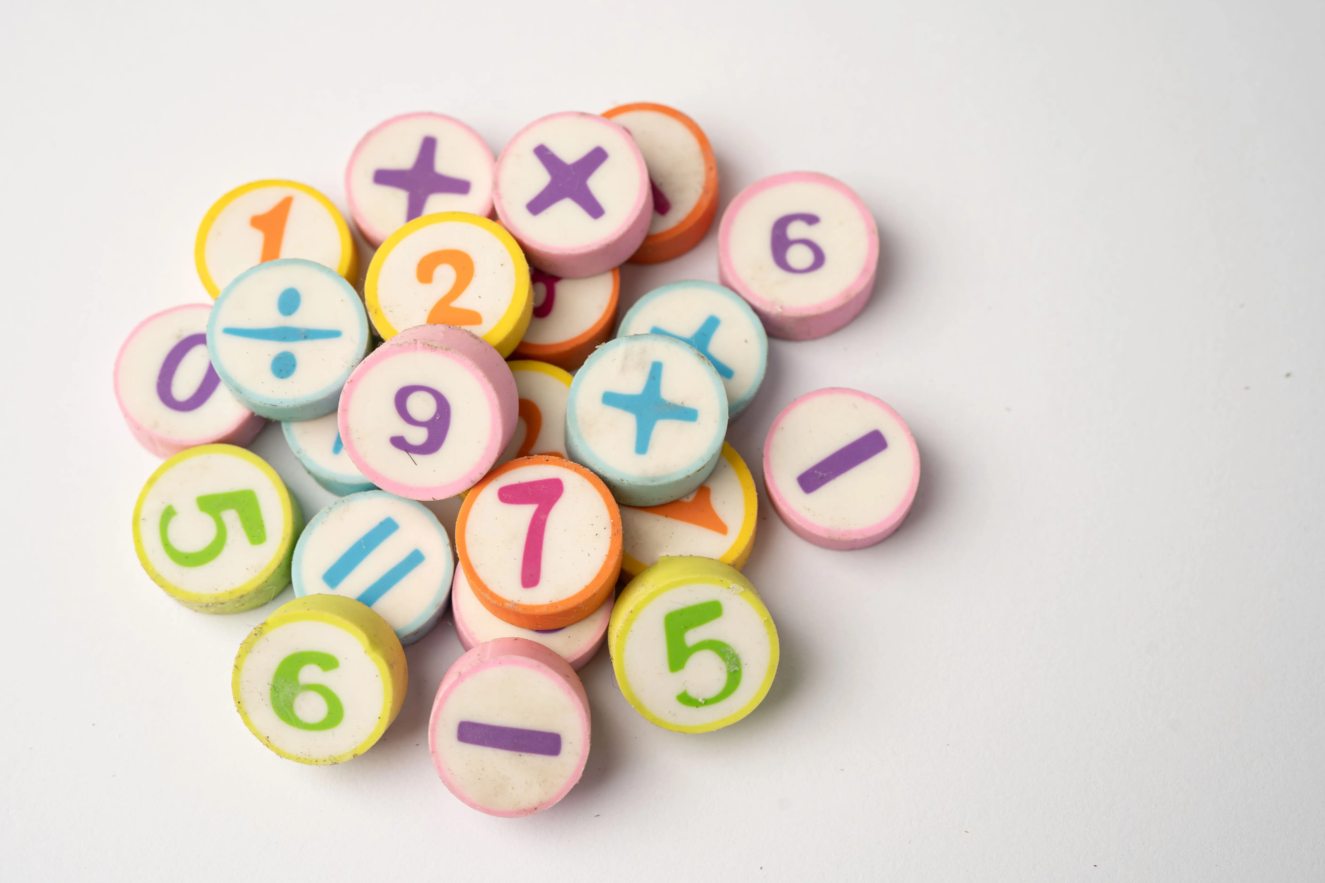 A pile of colorful round tokens displaying various mathematical symbols and numbers, including addition, subtraction, division, multiplication, and digits like 1, 2, 5, 6, 7, and 9. These tokens are placed on a white background, evoking a playful and educational vibe.