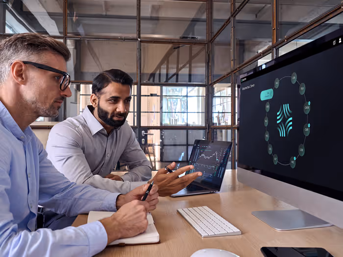 Two men discussing data charts displayed on a laptop and a large desktop monitor in a modern office.