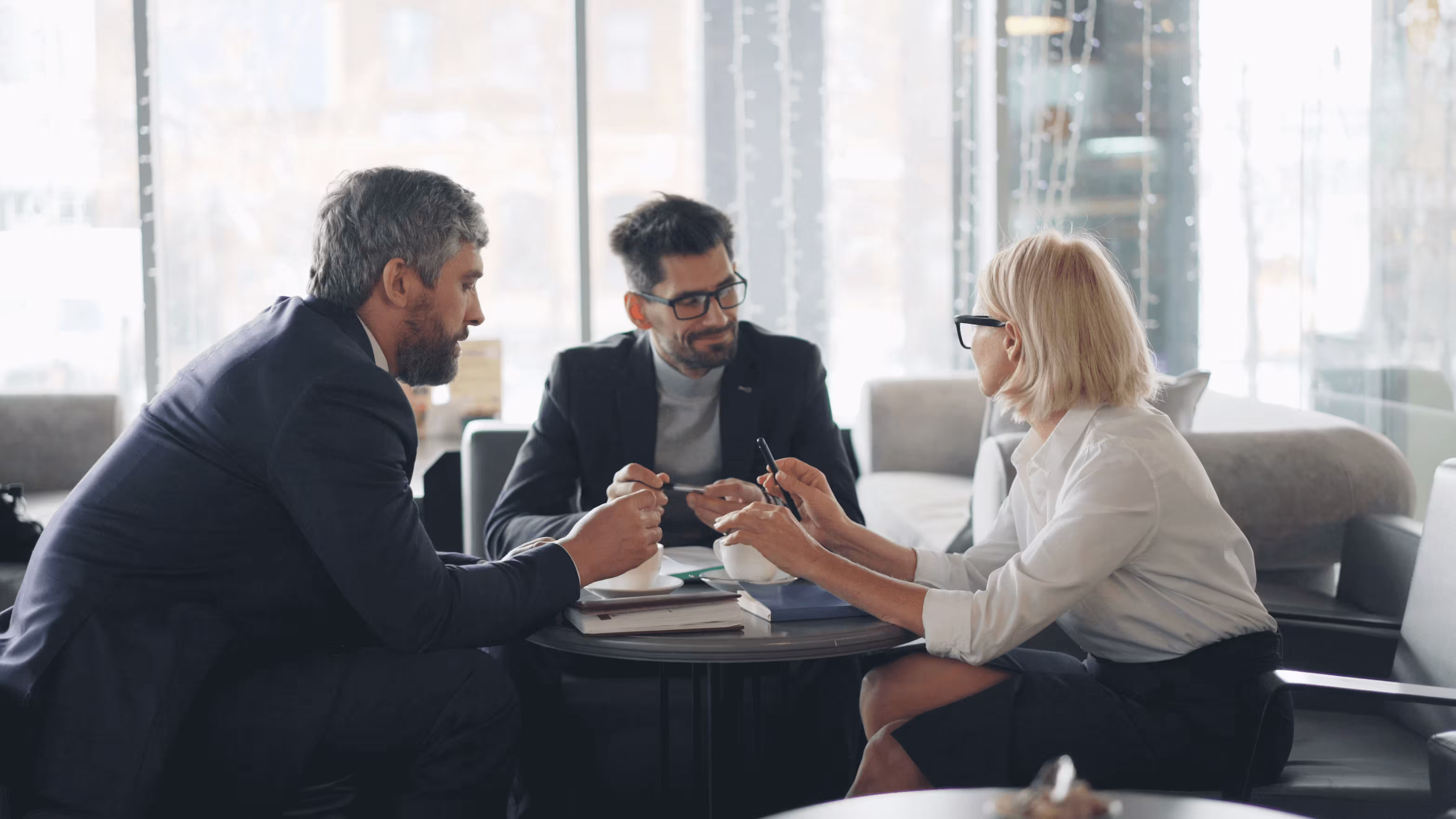 Three people sit around what is likely a work desk in an office. They seem to be discussing an idea amicably. They are wearing work clothes, there are big windows behind them - the environment is well lit by the sun light coming from them. 
