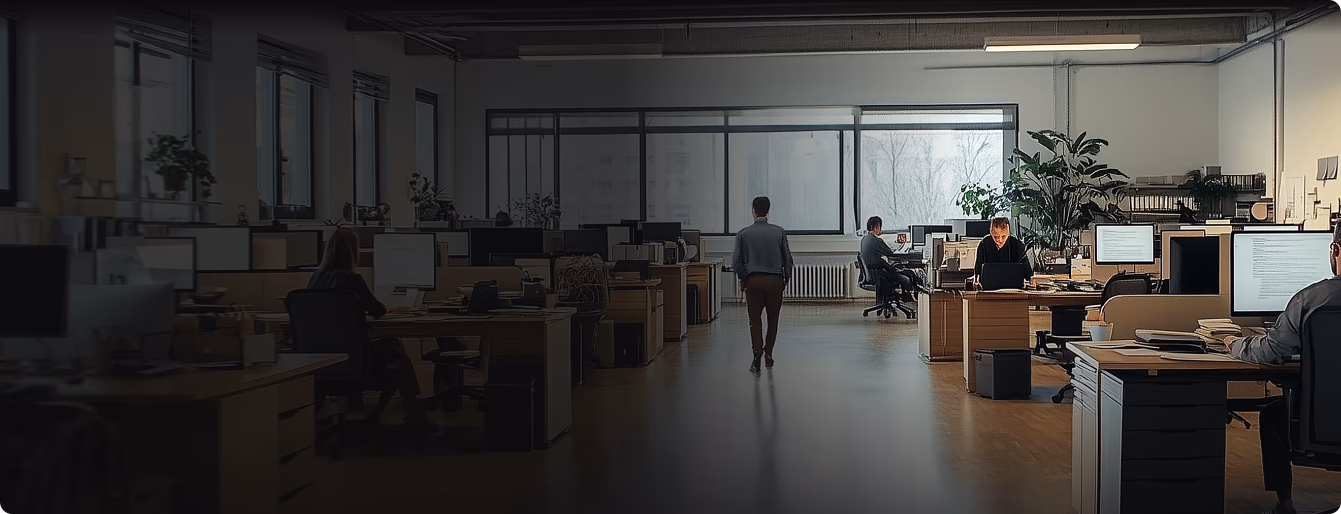 An office with wide windows, one person walks towards them. while others stare at their computer screens.