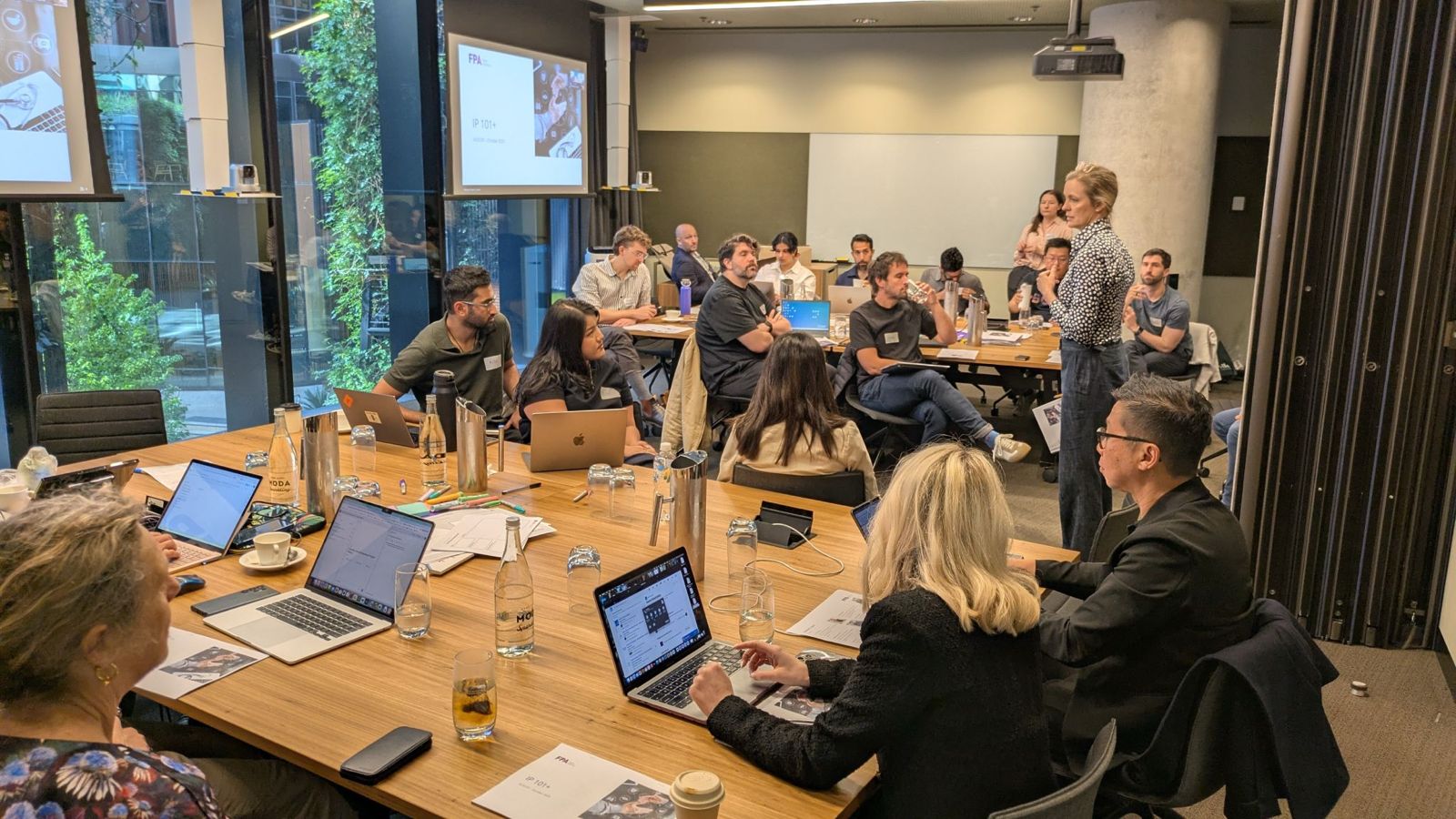A diverse group of people attending a meeting in a modern conference room with laptops and documents, listening to a woman standing and speaking.