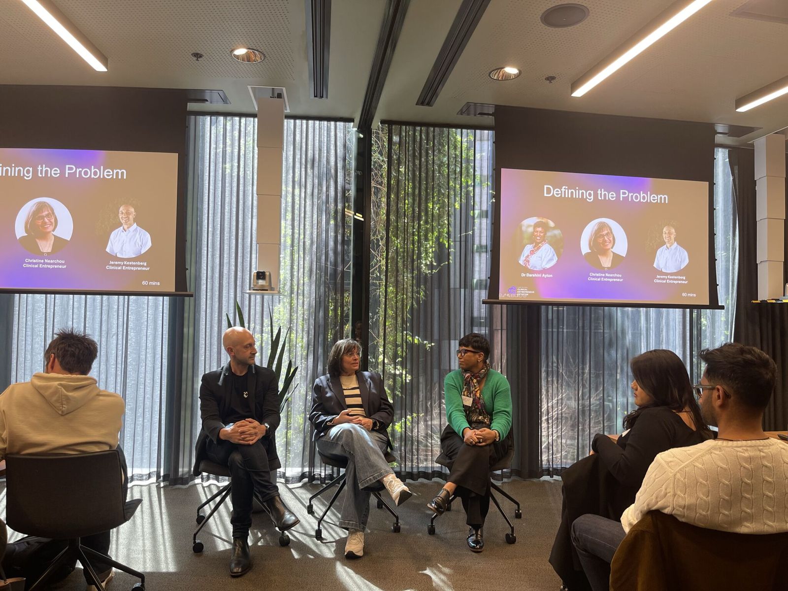 A panel discussion in a conference room with three panelists seated in front of large windows and two screens displaying 'Defining the Problem' with speaker photos and names.