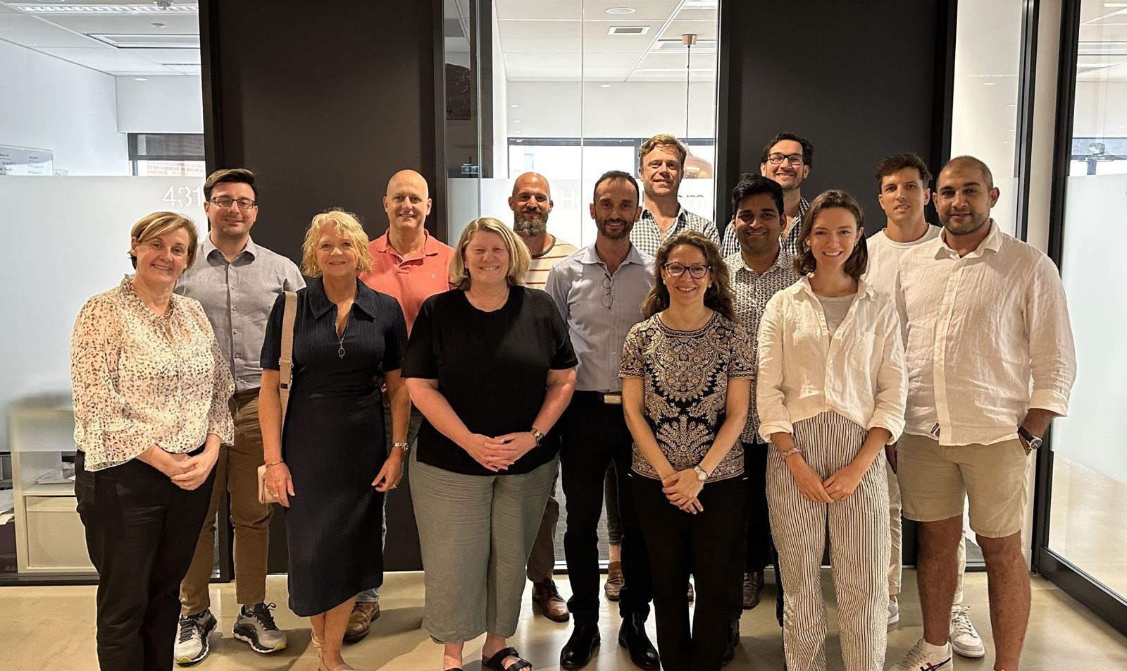 Group of fourteen people standing indoors in two rows, smiling at the camera.