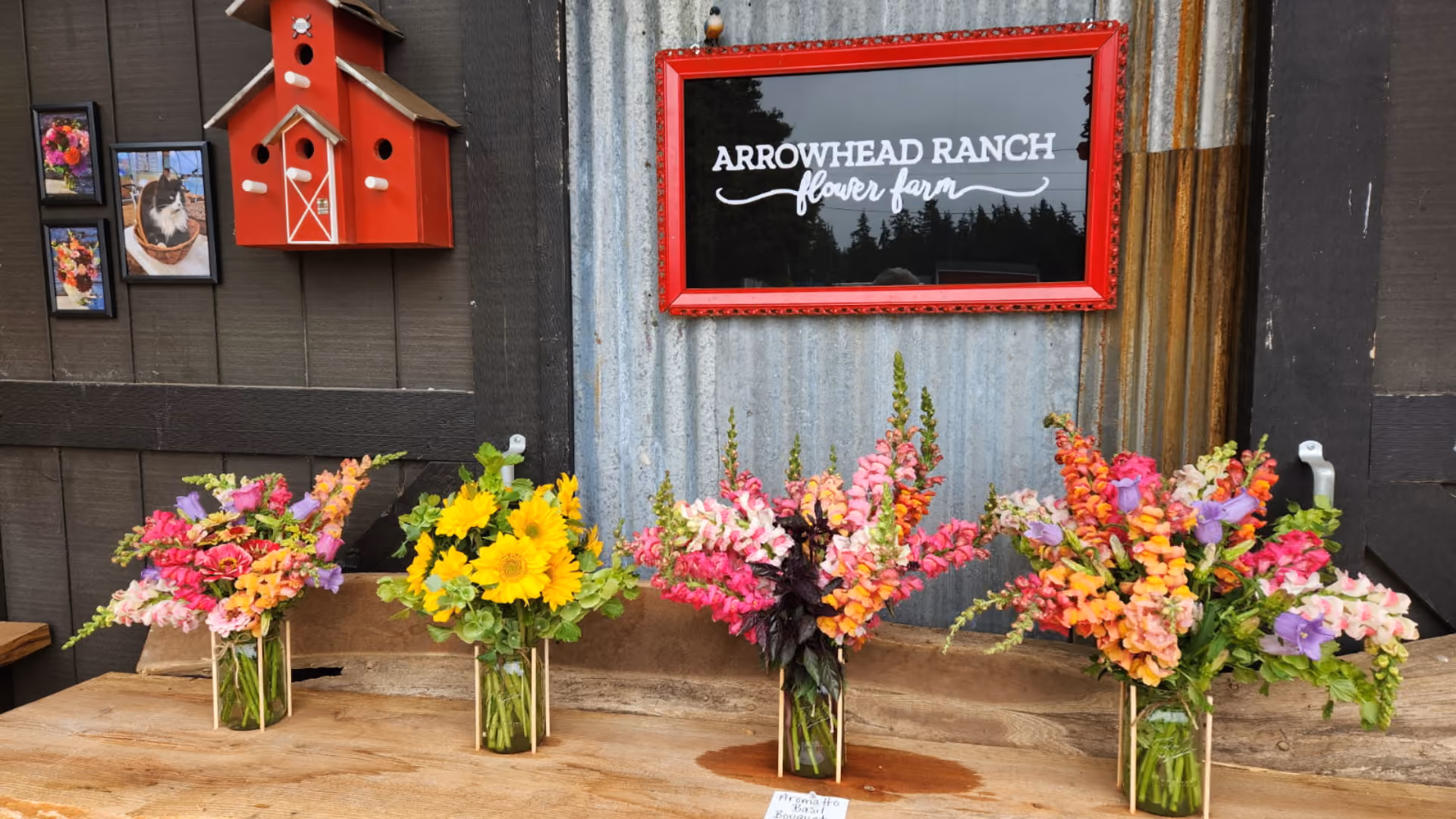 Four bouquets of fresh flowers in glass jars arranged on a wooden table under a sign that reads 'Arrowhead Ranch flower farm' with a rustic corrugated metal and wood background.