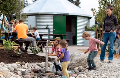 Children playing around a small water feature with rocks while adults watch nearby in an outdoor setting with a silo structure in the background.