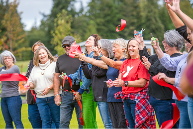 Group of people outdoors cheerfully cutting a red ribbon during a celebration event.