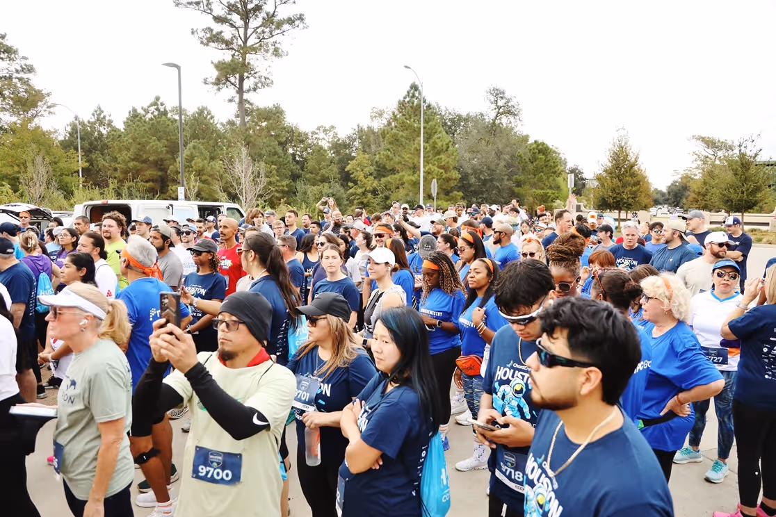 Large diverse group of runners gathered at the start line of an outdoor race event, many wearing blue Houston-themed shirts.