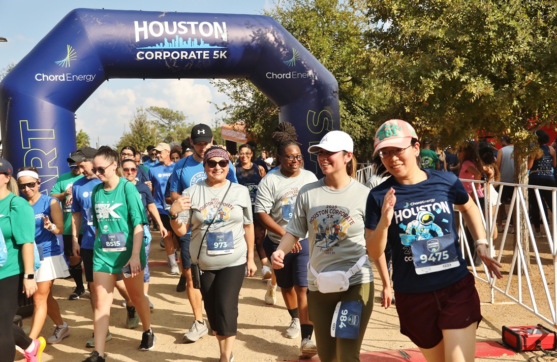 Diverse group of runners starting the Houston Corporate 5K race under a blue inflatable arch.