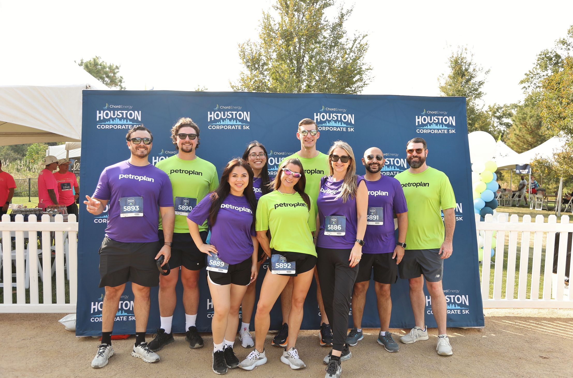 Group of nine runners wearing Petroplan shirts posing in front of a Houston Corporate 5K backdrop outdoors.