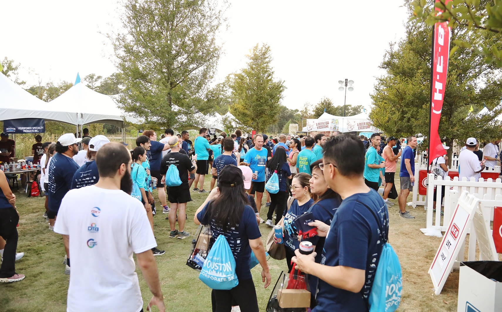 Crowd of people at an outdoor event with tents and trees, many wearing event shirts and carrying blue bags.