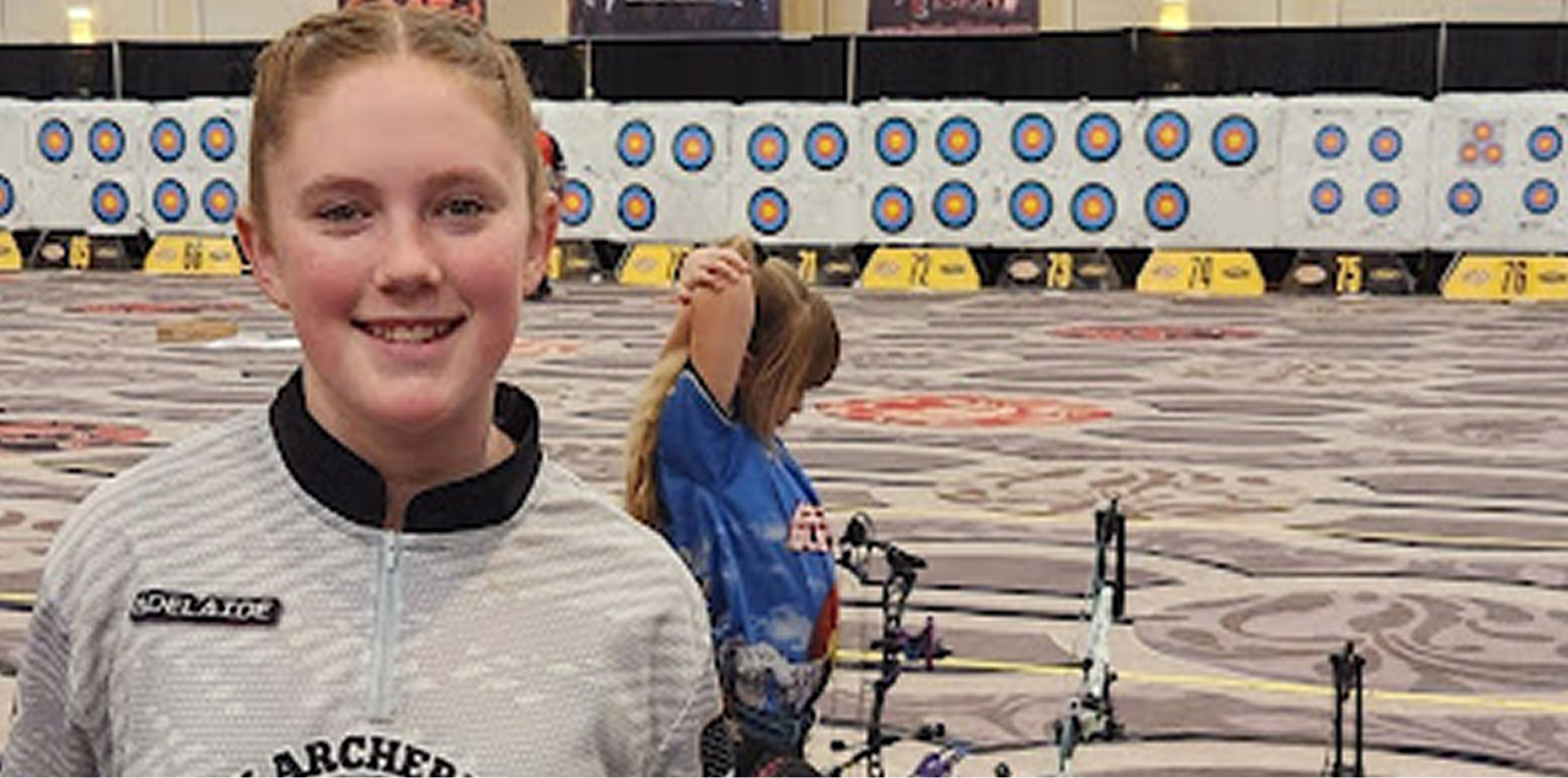 Image of young girl smiling and behind her is a person practicing archery