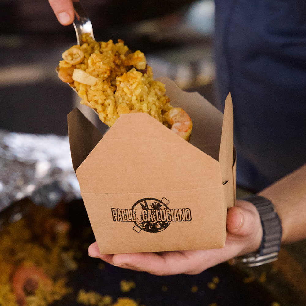 Hand serving a portion of paella in a take away box