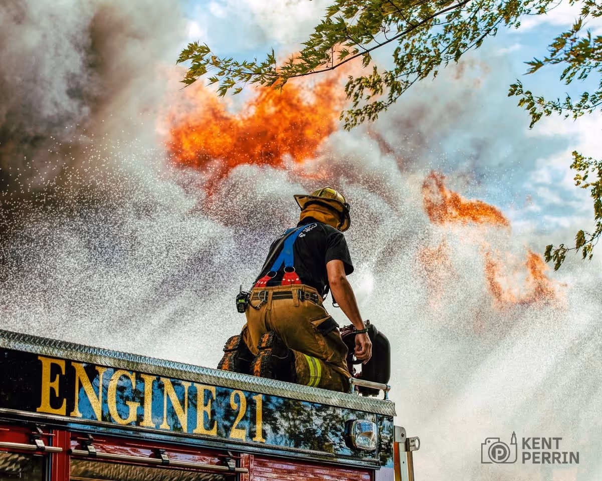 Firefighter on Engine 21 spraying water to extinguish large flames and smoke with tree branches overhead.