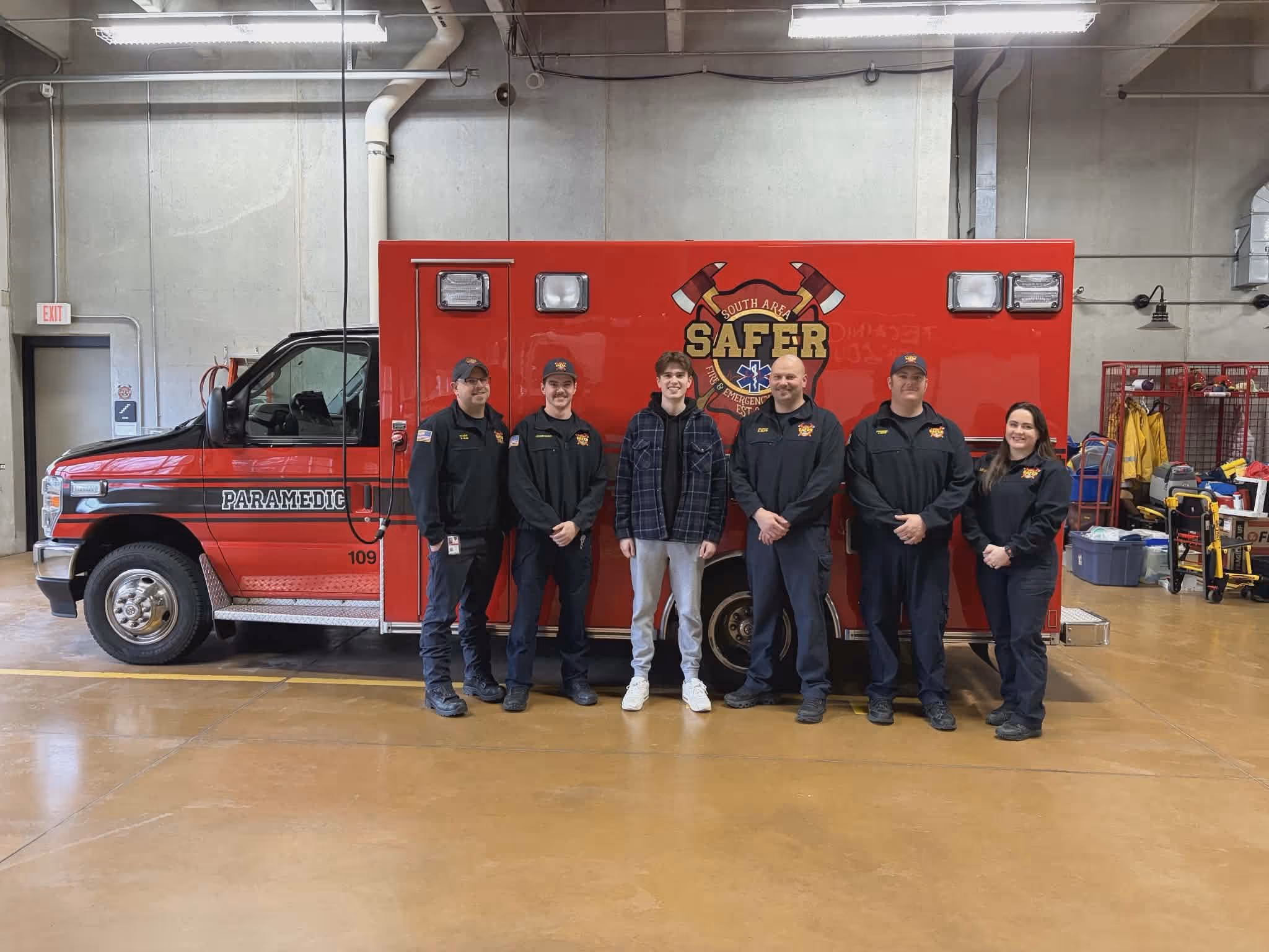 Five paramedics and one casually dressed young man standing in front of a red paramedic emergency vehicle inside a garage.