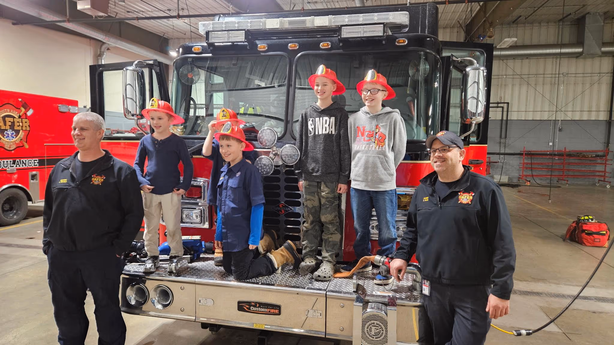 Two firefighters and five children wearing red plastic fire helmets posing in front of a fire truck inside a fire station.