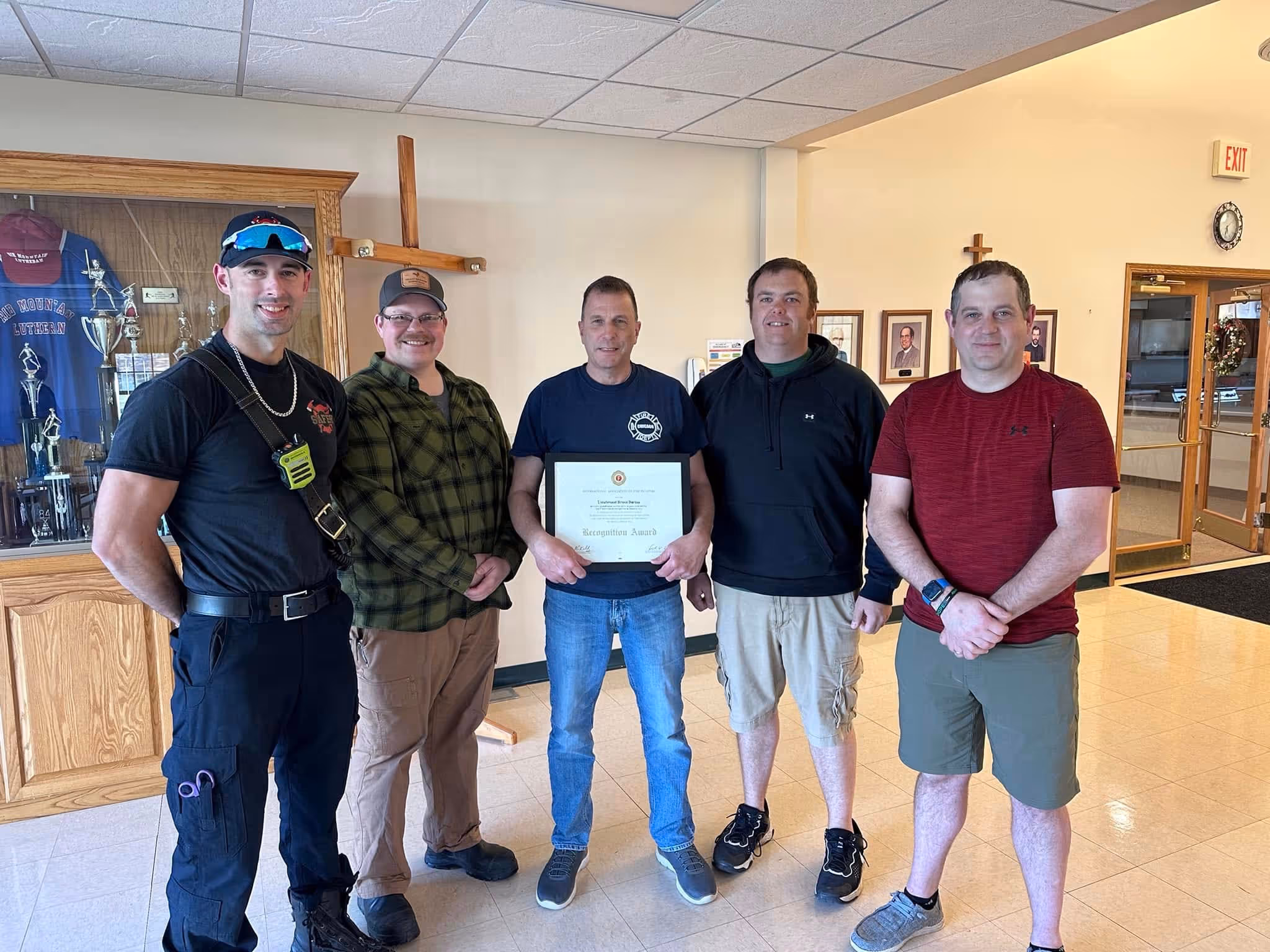 Group of five men standing indoors, one in center holding a framed recognition award.