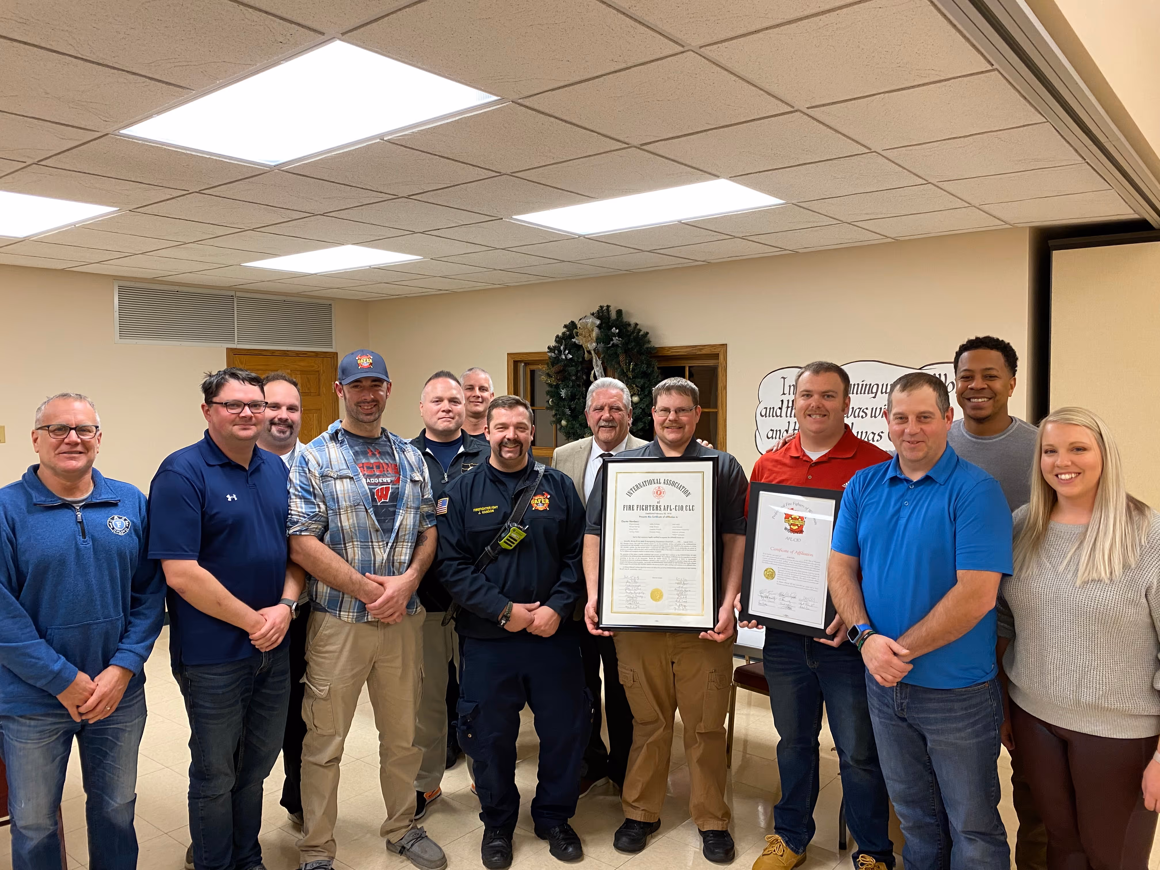 Group of smiling people standing indoors, some holding framed certificates, with a holiday wreath in the background.