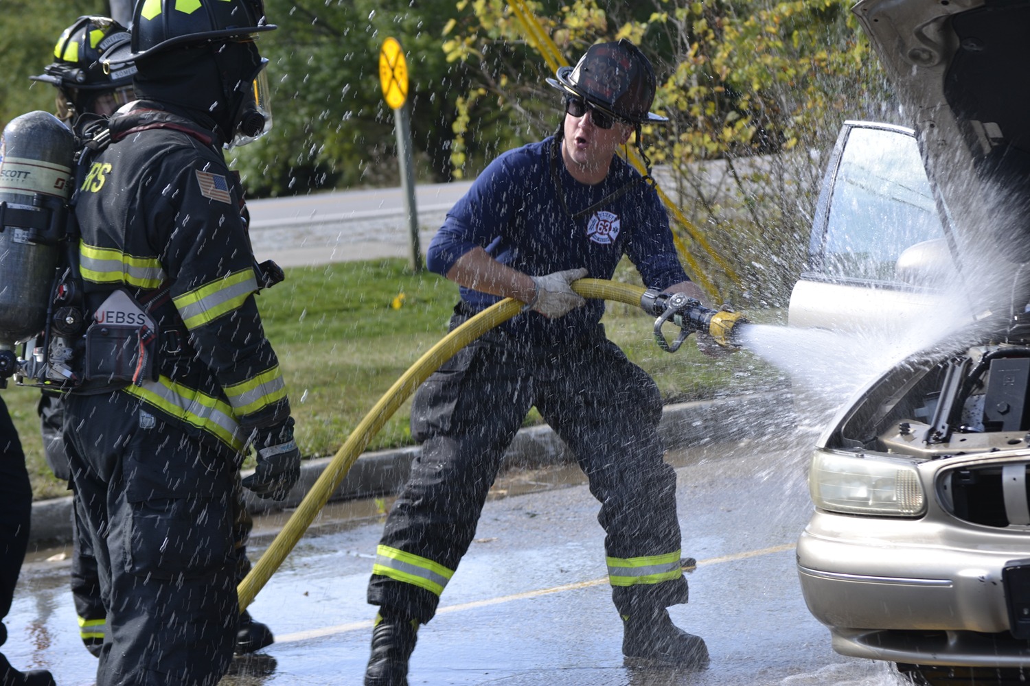 Two firefighters and five children wearing red plastic fire helmets posing in front of a fire truck inside a fire station.