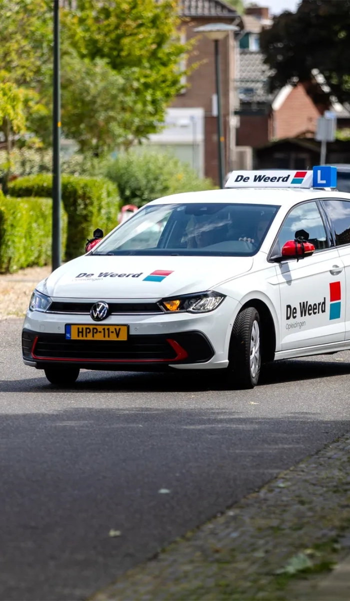 Witte lesauto van De Weerd Opleidingen met L-bord op het dak rijdt door een woonwijk
