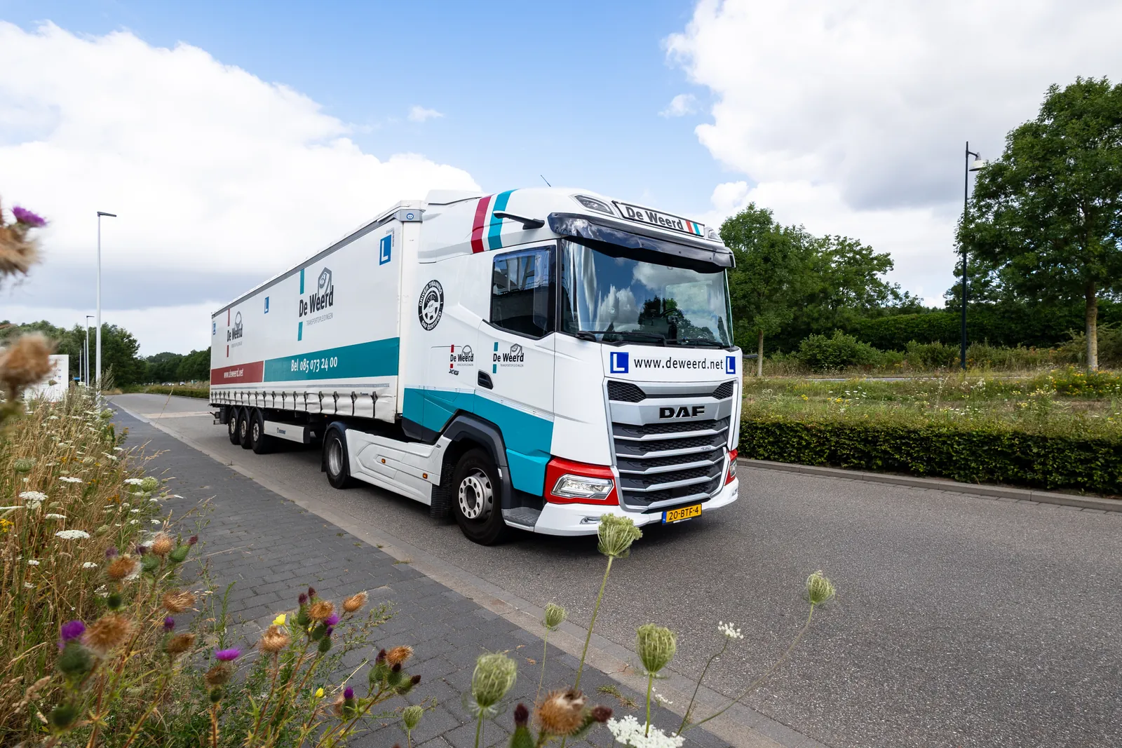 Lesvrachtwagen met oplegger van De Weerd Opleidingen met L-borden rijdt over een weg langs een berm met wilde bloemen