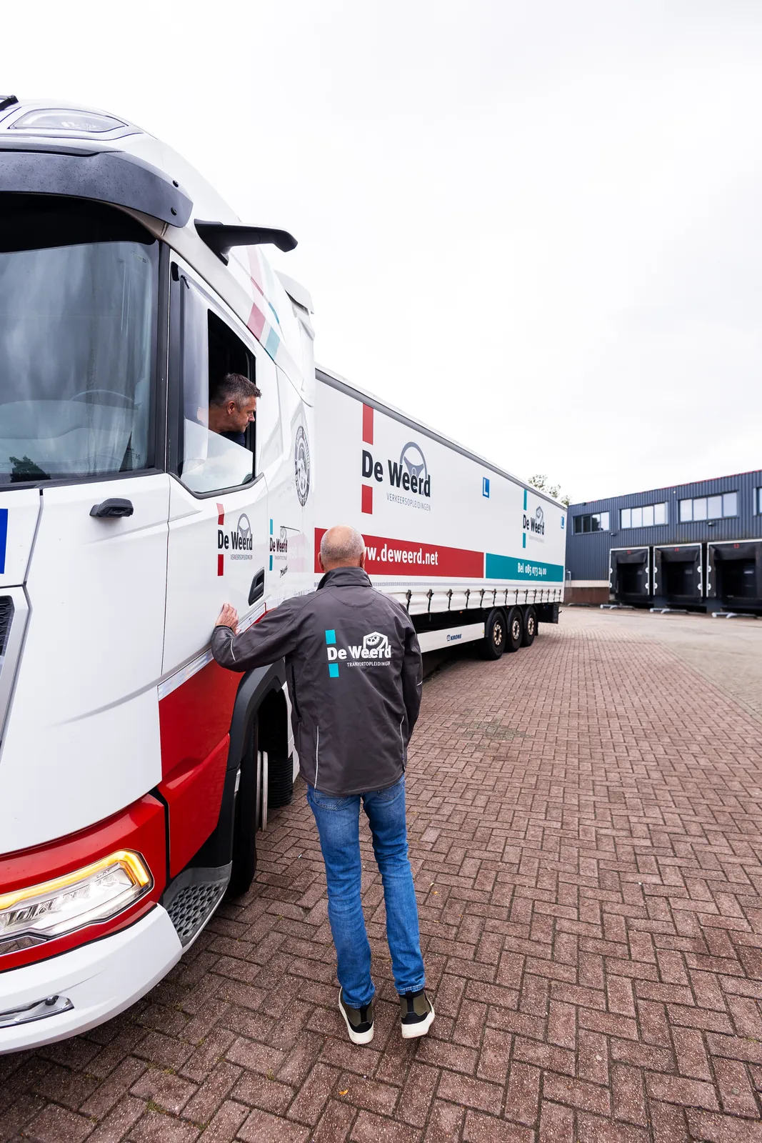 Instructeur van De Weerd Opleidingen staat naast de cabine van een vrachtwagen met oplegger en praat met de cursist achter het stuur